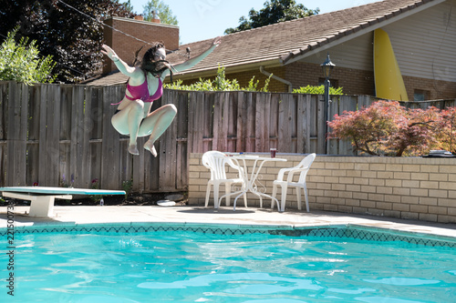 Young woman jumping off diving board into a backyard swimming pool arms up in the air legs tucked to her body caught in mid air. Girl in bikini jumps into outdoor swimming pool in the backyard.