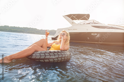 Красивая девушка плавает на надувном кругу, на заднем фоне плывет яхта, девушка пьет шампанское
