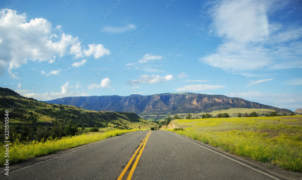 A highway dipping down between green hills dotted with trees with a mountain in the front in a sunny summertime Wyoming landscape