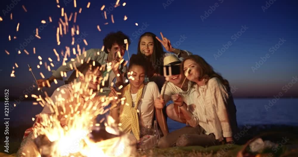 Five friends sitting near campfire during summer party, smiling, cheering, eating marshmallow and taking selfies on smartphone enjoying their time 4k Five friends sitting near campfire during summer party, smiling, cheering, eating marshmallow and taking selfies on smartphone enjoying their time 4k