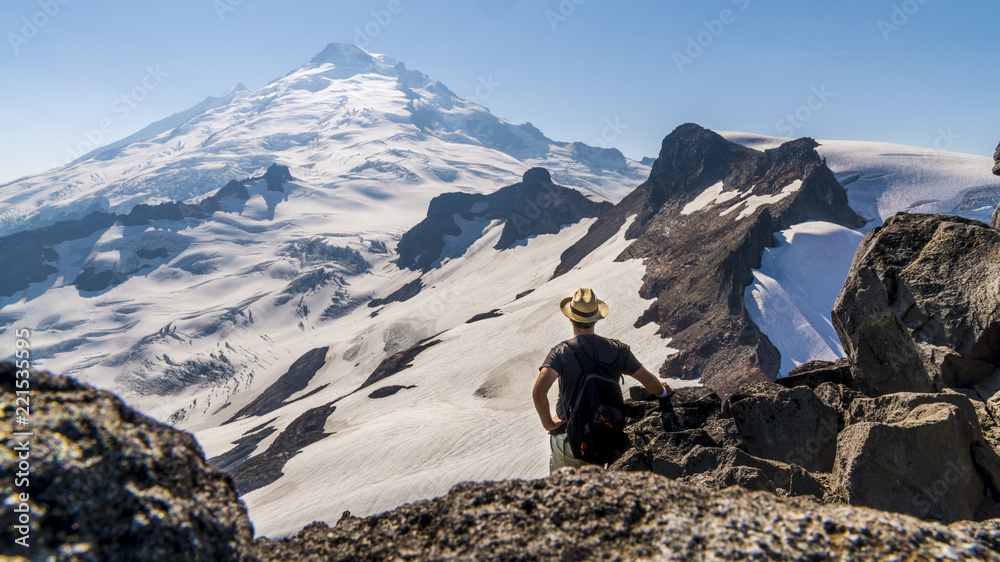 Beautiful panorama of a glacier, snowy mountain peak in the background, hiker wearing a backpack and a straw hat standing on the cliffs, admiring the beauty of nature. Mount Baker, Washington, USA.