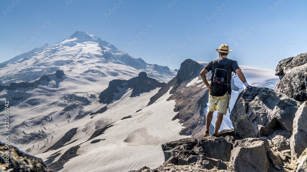 Beautiful panorama of a glaciar, snowy mountain peak in the background, hiker wearing a backpack and a straw hat standing on the cliffs, admiring the beauty of nature. Mount Baker, Washington, USA.