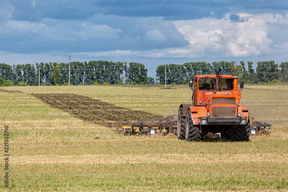 An orange modern tractor plows the earth in. a golden field of wheat on a summer day, in the sky a cumulus cloud, in the background a forest. An orange modern tractor plows the earth in. a golden field of wheat on a summer day, in the sky a cumulus cloud, in the background a forest.