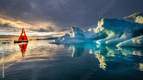 Greenland midnight sunset with heavy clouds mirror panorama with red sail ship and sun