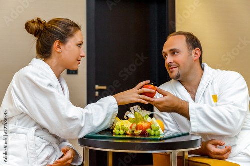 Young happy loving Caucasian couple of man and woman in white bathrobes talking after spa on honeymoon. Different fruits lying on the table. Man flirting to the woman and giving her an apple