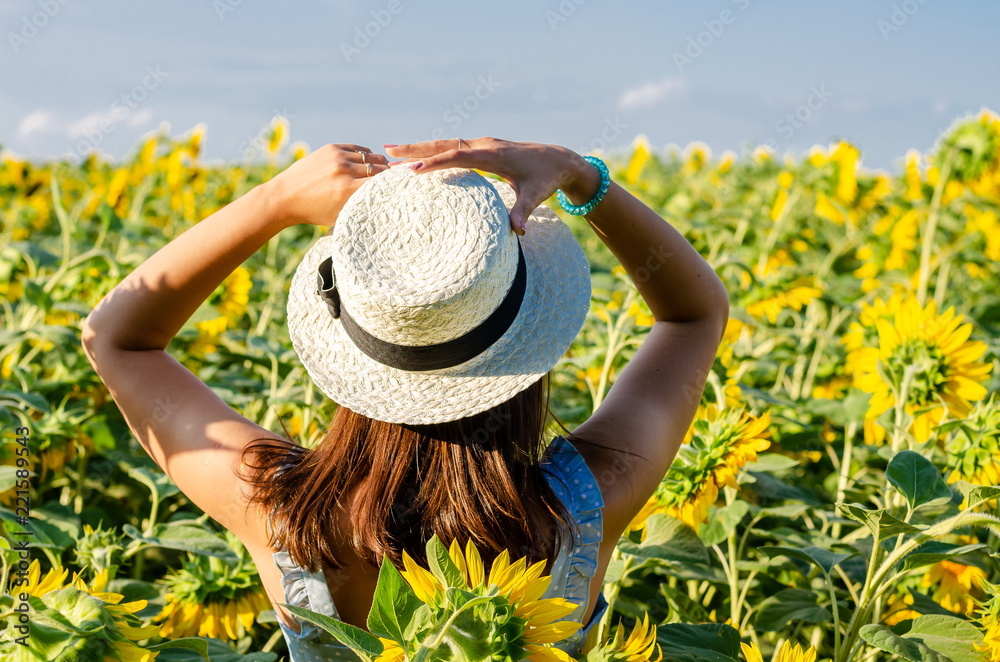 A girl in a hat stands with her back on a field of sunflowers and looks out into the distance to the horizon.