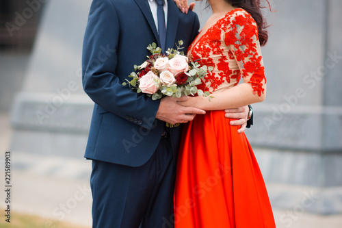The groom embraces his bride in a red wedding dress with a bouuet in hands