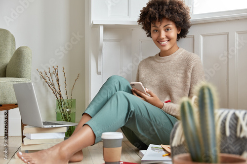 Photo of satisfied marketing expert dressed in casual clothes, enjoys cozy atmosphere, smiles, surrounded with pile of books and electronic gadget, updates new application, develops website design