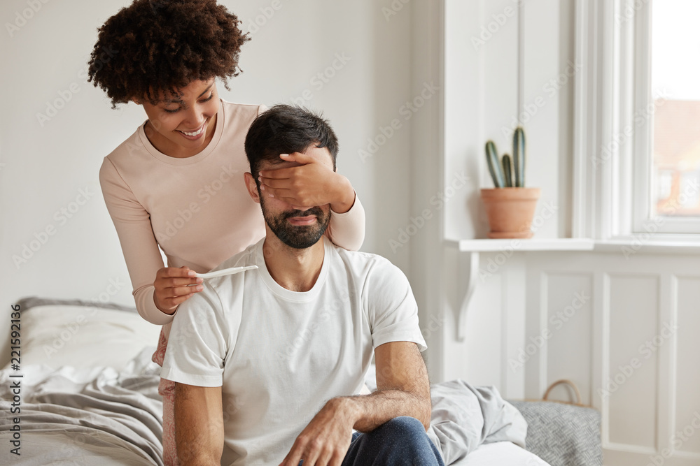 Φωτογραφία Indoor shot of happy wife makes surprise to husband, covers his eyes and holds pregnancy test, informs he will become father going to share good news, pose together in bedroom