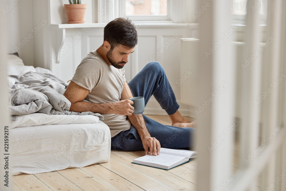 Relaxed bearded Caucasian guy enjoys spare time, calm atmosphere at home, holds mug with coffee, reads interesting novel, looks attentively at book, sits on floor near not made bed. Hobby concept
