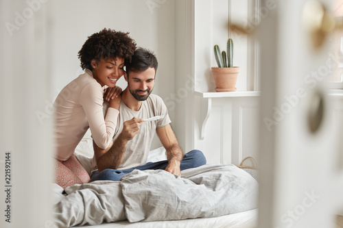 Mixed race family couple rejoice good news in morning, find out about pregnancy, sit together on bed and look happily at control line, have nice relationships. Motherhood and fatherhood concept