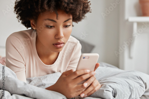 Cropped image of dark skinned cute teenage girl dressed in casual clothes, enjoys Sunday morning in bed, holds cell phone, has online communication, free internet connection. Leisure concept