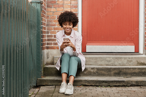Street style concept. Beautiful curly dark skinned hipster enjoys spare time, sits on threshold of old house, waits for friend outdoor, enjoys online communication via cell phone, connected to wifi
