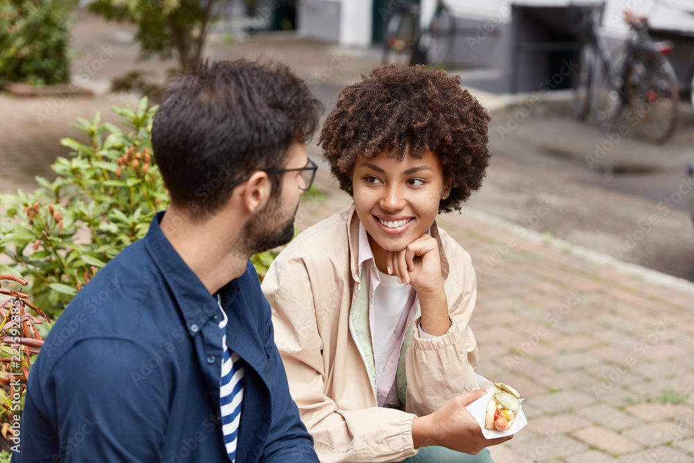 Fototapeta premium Happy mixed race couple pose in urban setting, have pleasant talk together, eat hot dog, enjoy spending free time in unknown city. Tourists have stroll at street. Lifestyle and friendship concept