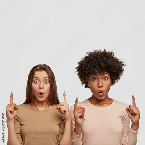 Isolated shot of mixed race women with surprised expression notice plane in sky, stand closely to each other, isolated over white background with copy space for your promotional content. Diversirty