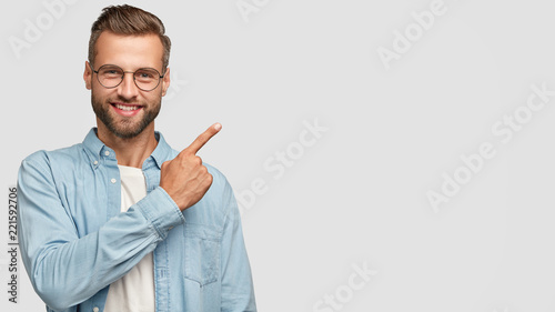 This idea just fascinated me. Attractive young unshaven individual with happy expression, indicates with index finger at blank copy space, advertises you place to vist, isolated over white wall