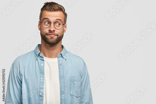 Isolated shot of funny bearded man designer or freelancer looks thoughtfully aside, thinks on how climb career ladder, wears casual shirt and round spectacles, isolated on white wall with copy space photo