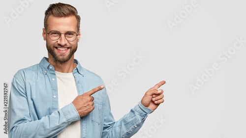 Positive unshaven man looks and points at upper right corner with both index fingers, smiles with approval, suggest going there, sees something positive and very interesting, isolated on white wall