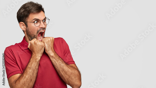 Crazy man with terrific expression keeps mouth widely opened, exclaims furiously, being puzzled by something, dressed in casual outfit, isolated over white background with copy space for your text photo