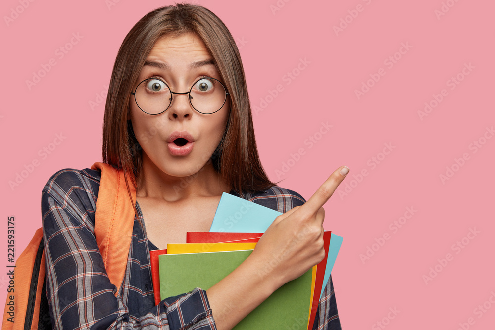 Horizontal shot of stupefied brunette pupil has surprised scared expression, points at upper right corner with index finger, dressed in checkered shirt, carries dictionary, going in school library.
