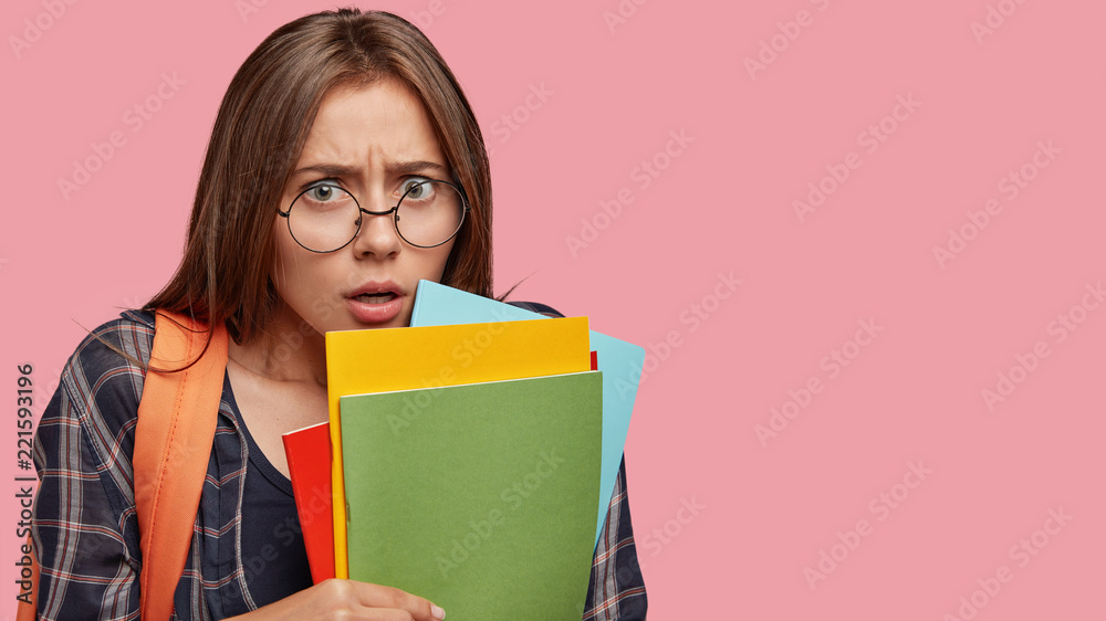 Horizontal shot of scared brunette woman with puzzled expression, frowns face, holds many books, feels nervous before answering during class, stands against pink studio wall with blank space Horizontal shot of scared brunette woman with puzzled expression, frowns face, holds many books, feels nervous before answering during class, stands against pink studio wall with blank space