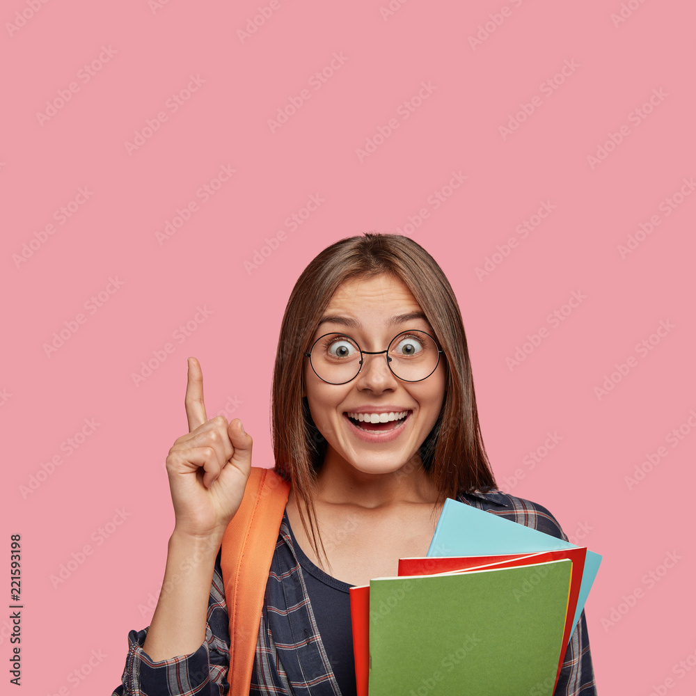 Vertical photo of amazed lovely woman holds textbooks, points with index finger upwards, shows blank space above head, being in high spirit, isolated over pink background. People, reading, learning Vertical photo of amazed lovely woman holds textbooks, points with index finger upwards, shows blank space above head, being in high spirit, isolated over pink background. People, reading, learning