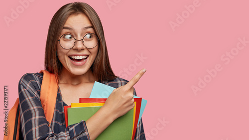Indoor shot of pretty happy schoolgirl attracts your attention at copy space, advises use it wisely, dressed in casual outfit, holds textbooks going home after university or college isolated on pink
