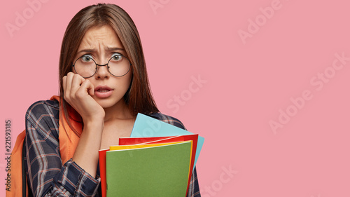 Nervous stressful student worries before presenting diploma paper in auditorium, keeps hand on cheek, holds books, carries rucksack, isolated over pink wall with blank space for your text or promotion