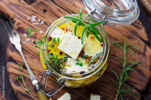 Feta cheese marinated in olive oil with fresh herbs in glass jar. Wooden background. Top view.