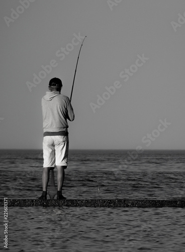 Vertical black and white photo of a lone fisherman standing on a concrete pier in the sea. 