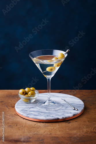 Martini cocktail with green olives on marble board. Wooden table. Blue background.