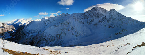 Scenic panoramic winter landscape of snowy Caucasus mountain range with peaks and clouds on blue sky sunny day. Elbrus, Cheget, Baksan