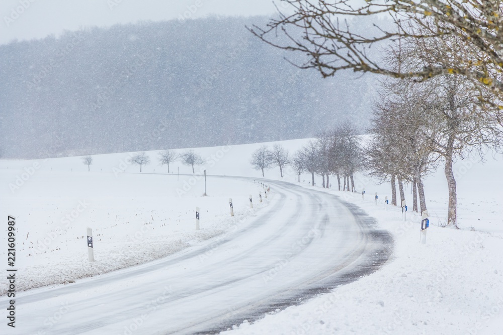 Fototapeta premium Leere glatte Winter Straße mit Schnee wartet auf Winterdienst. Die Fahrbahn ist weiß glatt verschneit. Vorsicht vor Unfall durch Schneesturm und schlechte Sicht. Wetter zur Weihnachten Jahreszeit