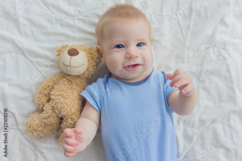 the baby lies on the floor with a teddy bear. A child waving his hand while lying on the floor.