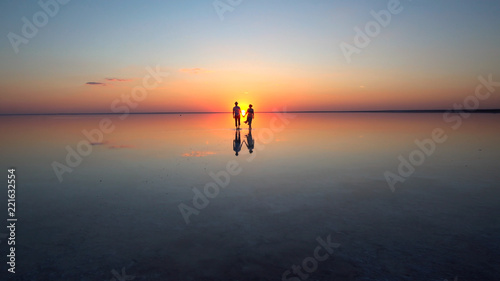 A young couple walking on calm mirrored surface of shallow water into the setting sun.. They are holding each other s hands