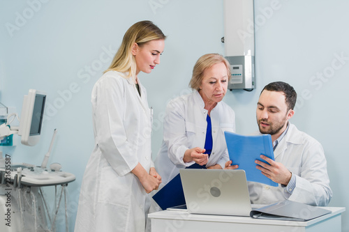 Photo of three doctors together discussing new way of treatment while having a meeting at office. Doctors using laptop computer while discussing new way of treatment.