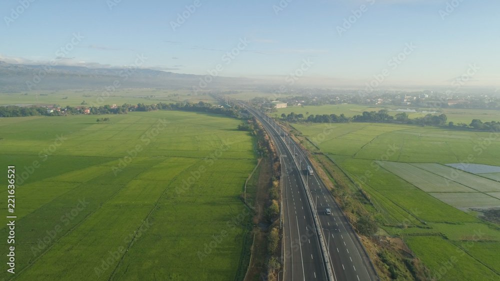 Aerial view highway with cars among farmer fields, rice terraces on background of mountains. Philippines, Luzon. High speed highwayin the morning sunrise. Tropical landscape in Asia.