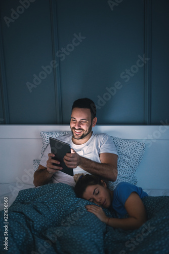 Handsome beautiful young man sitting in a bed and holding a tablet while his girlfriend is lying on his legs.