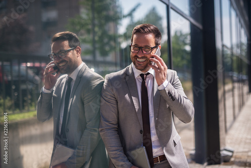 Lucky day at work.Smiling confident businessman talking with his colleague about successful ending job. Standing outside leaned on building wall.