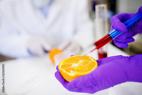 Close up of a hand in a protective glove holding orange and taking a sample from it with a syringe.