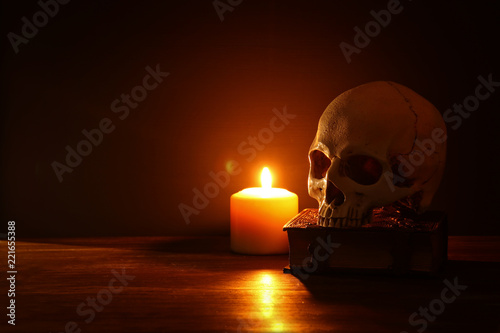 Human skull, old book and burning candle over old wooden table and dark background.