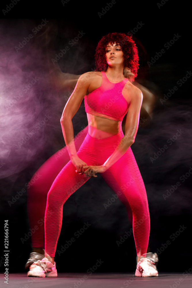Happy young and fitness woman dancing on black background with smoke. Mixed lighting photography.