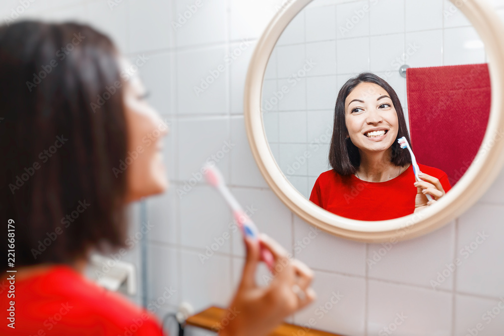Asian woman brushing teeth and looking in the mirror in the bathroom in her apartment