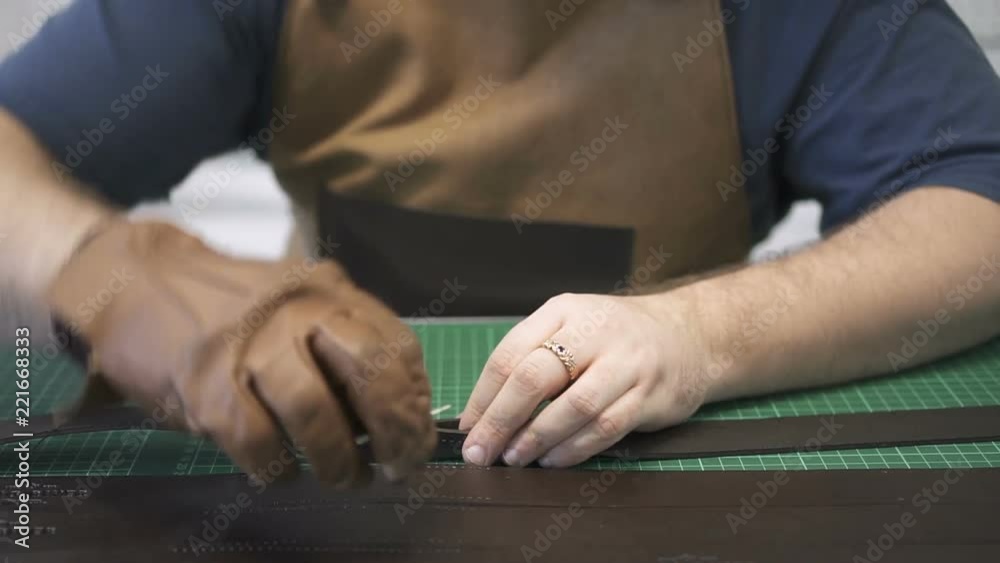 American entrepreneur works with real leather at desk in lighting room, front view of young specialist wearing uniform sews new product for import, sitting in workspace in modern interior. Concept American entrepreneur works with real leather at desk in lighting room, front view of young specialist wearing uniform sews new product for import, sitting in workspace in modern interior. Concept