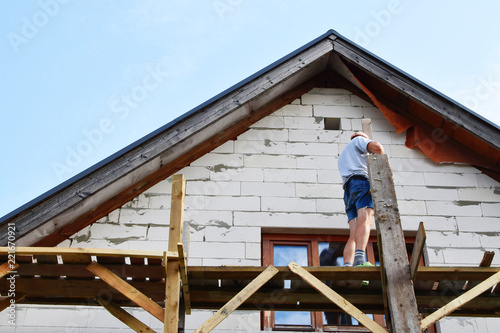 Wallpaper Mural Man installing wooden planks and works on wooden scaffolding next to unfinished lightweight concrete block house under construction in summer day. Home improvement concept. Torontodigital.ca