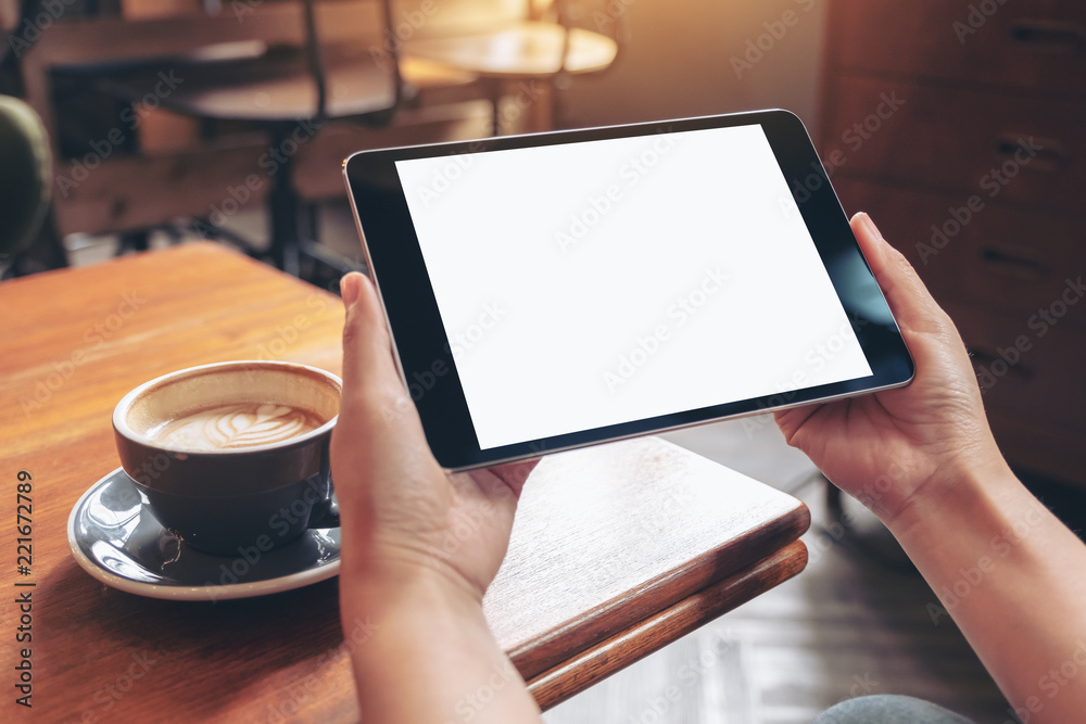 Mockup image of hands holding black tablet pc with blank white screen with coffee cup on wooden table in cafe Mockup image of hands holding black tablet pc with blank white screen with coffee cup on wooden table in cafe