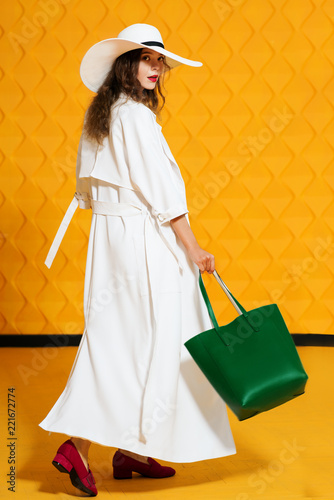 Full-length studio fashion portrait of young beautiful model wearing long white trench coat, wide-brimmed hat, pink loafers, holding green faux-leather shopper bag, posing on yellow background 