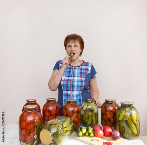 woman in blue plaid tunic in the kitchen next to the table. on the table are jars of pickled, salted tomatoes and cucumbers, on the table are fresh tomatoes, cucumbers, onions, garlic, vegetables photo