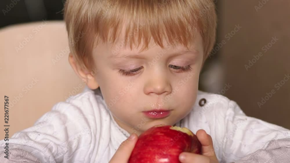Close-up of a small blond boy eating a red apple sitting at a table at home, high resolution. Cute little child is biting red apple while sitting at table. Close-up of a small blond boy eating a red apple sitting at a table at home, high resolution. Cute little child is biting red apple while sitting at table.
