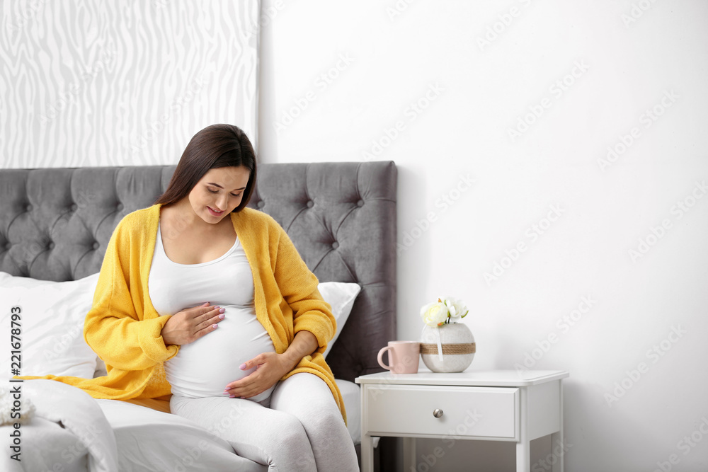 Young beautiful pregnant woman sitting on bed and touching her belly at home